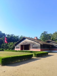 
A wooden building with a thatched roof, set against a clear blue sky. In front of the building, there is a neatly manicured lawn with low hedges, and a red nepali flag is prominently displayed. 