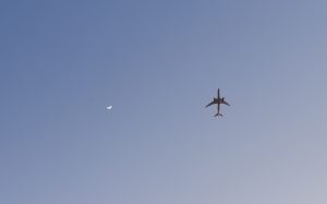 A commercial airplane, viewed from below, flies across a clear blue daytime sky near a thin crescent moon.