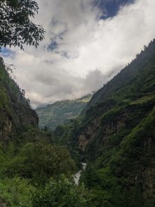 A scenic valley landscape featuring steep green mountainsides, a river flowing through the center, and dramatic clouds overhead with a visible tiny helicopter in the sky.