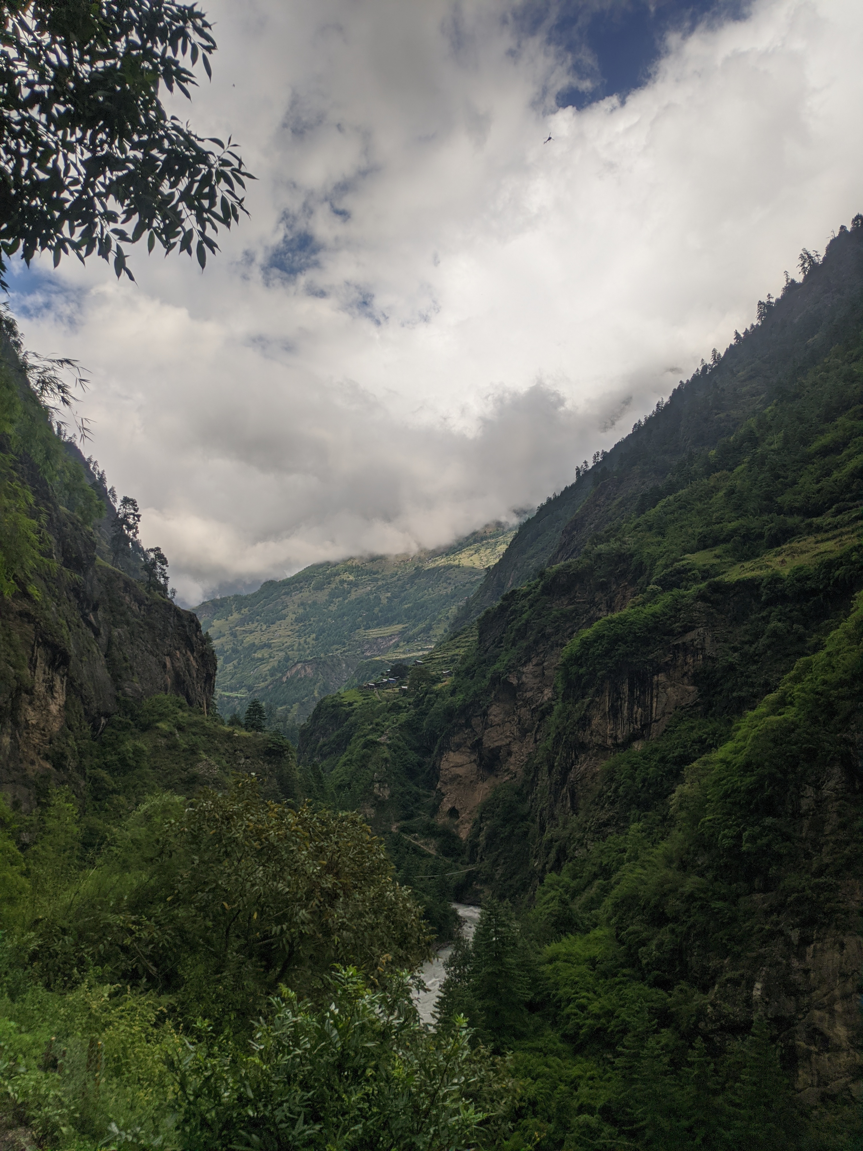 A scenic valley landscape featuring steep green mountainsides, a river flowing through the center, and dramatic clouds overhead with a visible tiny helicopter in the sky.