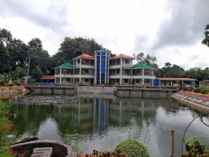 A large, colorful building with red and green roofs is reflected in a serene pond. Surrounding greenery and cloudy skies create a calm atmosphere.