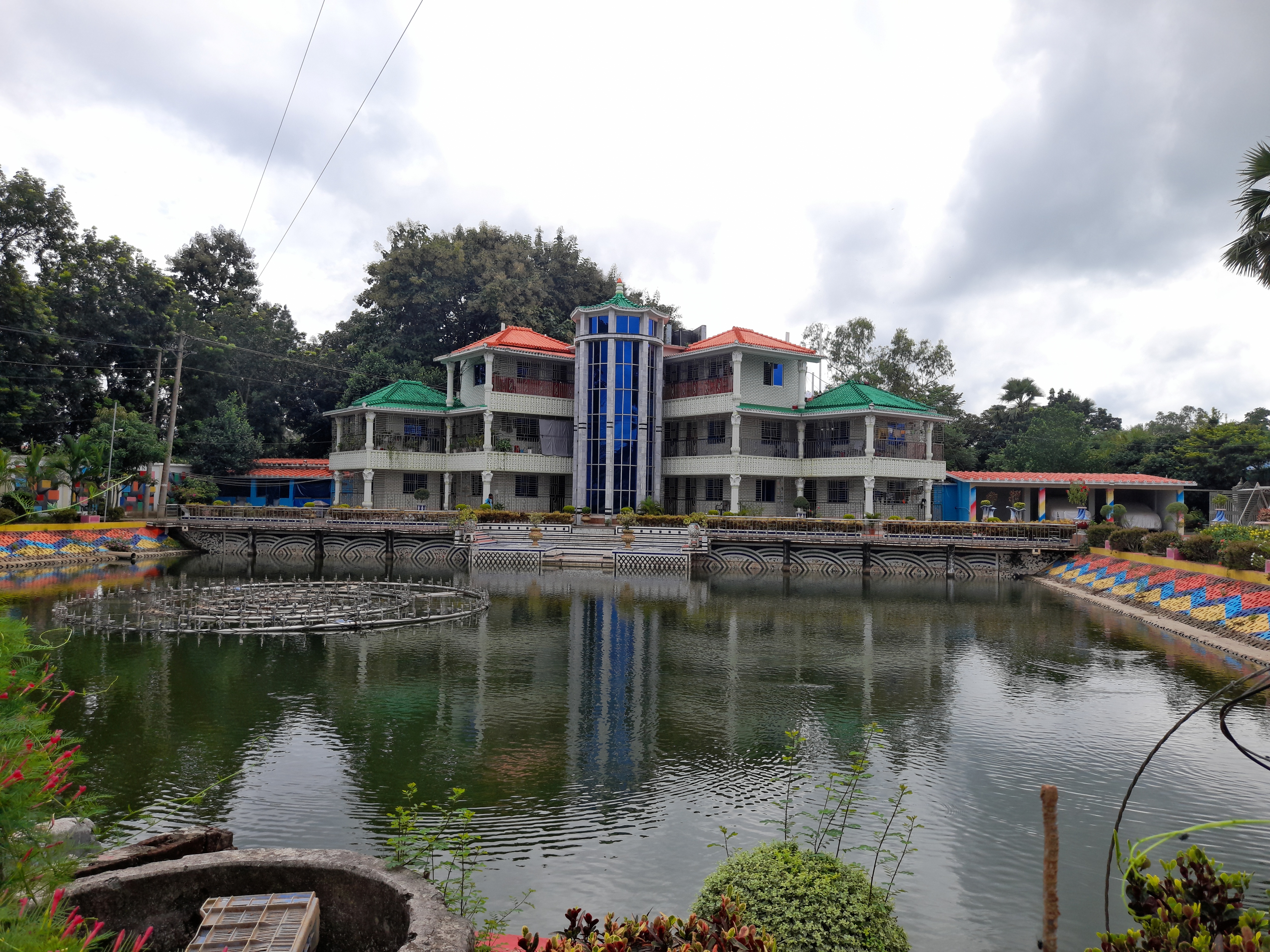 A large, colorful building with red and green roofs is reflected in a serene pond. Surrounding greenery and cloudy skies create a calm atmosphere.
