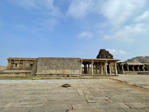 A wide-angle view of the Shree Vijay Vitthala Temple in Hampi, Karnataka, featuring ancient stone walls and pillared corridors under a blue sky. This shot captures the grandeur and symmetry of the temple’s outer structure. 