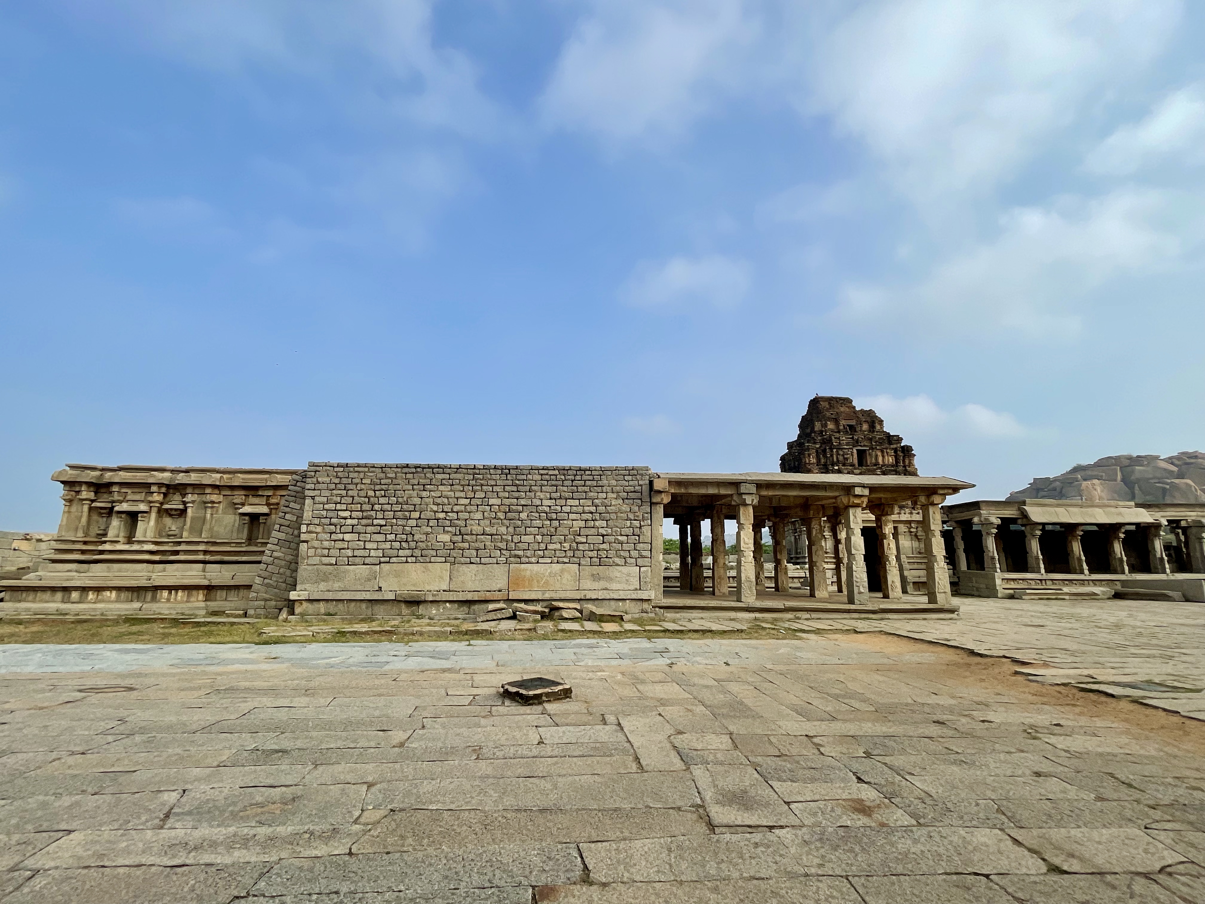 A wide-angle view of the Shree Vijay Vitthala Temple in Hampi, Karnataka, featuring ancient stone walls and pillared corridors under a blue sky. This shot captures the grandeur and symmetry of the temple’s outer structure.