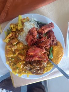 A colorful plate with yellow rice, mixed vegetables, crispy fried chicken, creamy sauce, meat in gravy, and a small serving of okra on a light tablecloth.