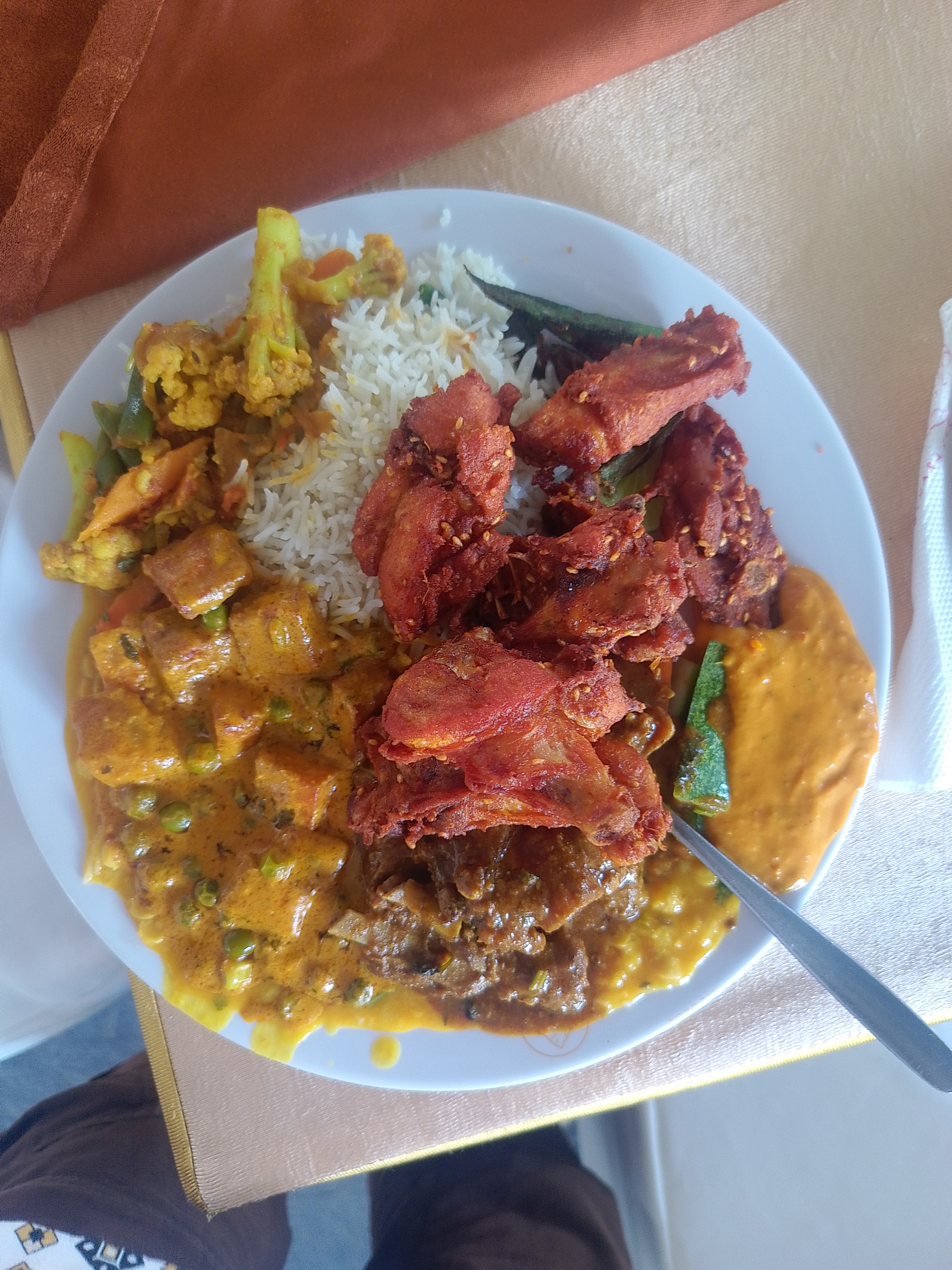 A colorful plate with yellow rice, mixed vegetables, crispy fried chicken, creamy sauce, meat in gravy, and a small serving of okra on a light tablecloth.