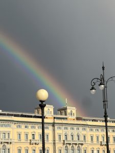 A bright rainbow arches across a dark, stormy sky above a long, yellow building with classic architecture. Two decorative streetlights frame the foreground.