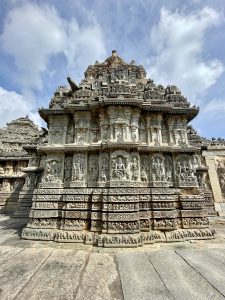 The outer wall of the Lakshmi Narasimha Temple in Nuggehalli, Hassan, is beautifully adorned with detailed sculptures of deities and motifs under clear blue skies. 