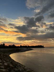 A tranquil waterscape at sunset featuring a curving shoreline lined with smooth stones. 