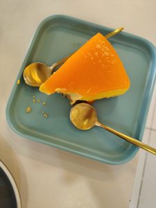 A slice of bright orange gelatin cake sits on a light blue square plate, accompanied by two golden spoons.