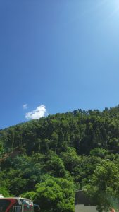 A bright blue sky with a few scattered clouds above lush green hills, densely covered with trees.