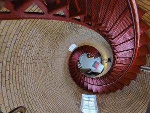 The interior view of a spiral staircase looking downward from the top inside a lighthouse. The staircase is made of metal, painted red, while the surrounding structure is made of light-colored bricks. 
