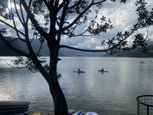 Fewa Lake reflects clouds and mountains, framed by tree branches, with kayakers in the distance and colorful paddleboards nearby.