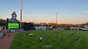 A baseball field, shaded by evening. There are people all over the field playing lawn games.  There's a mascot visible on the field as well. Grand Rapids, Michigan