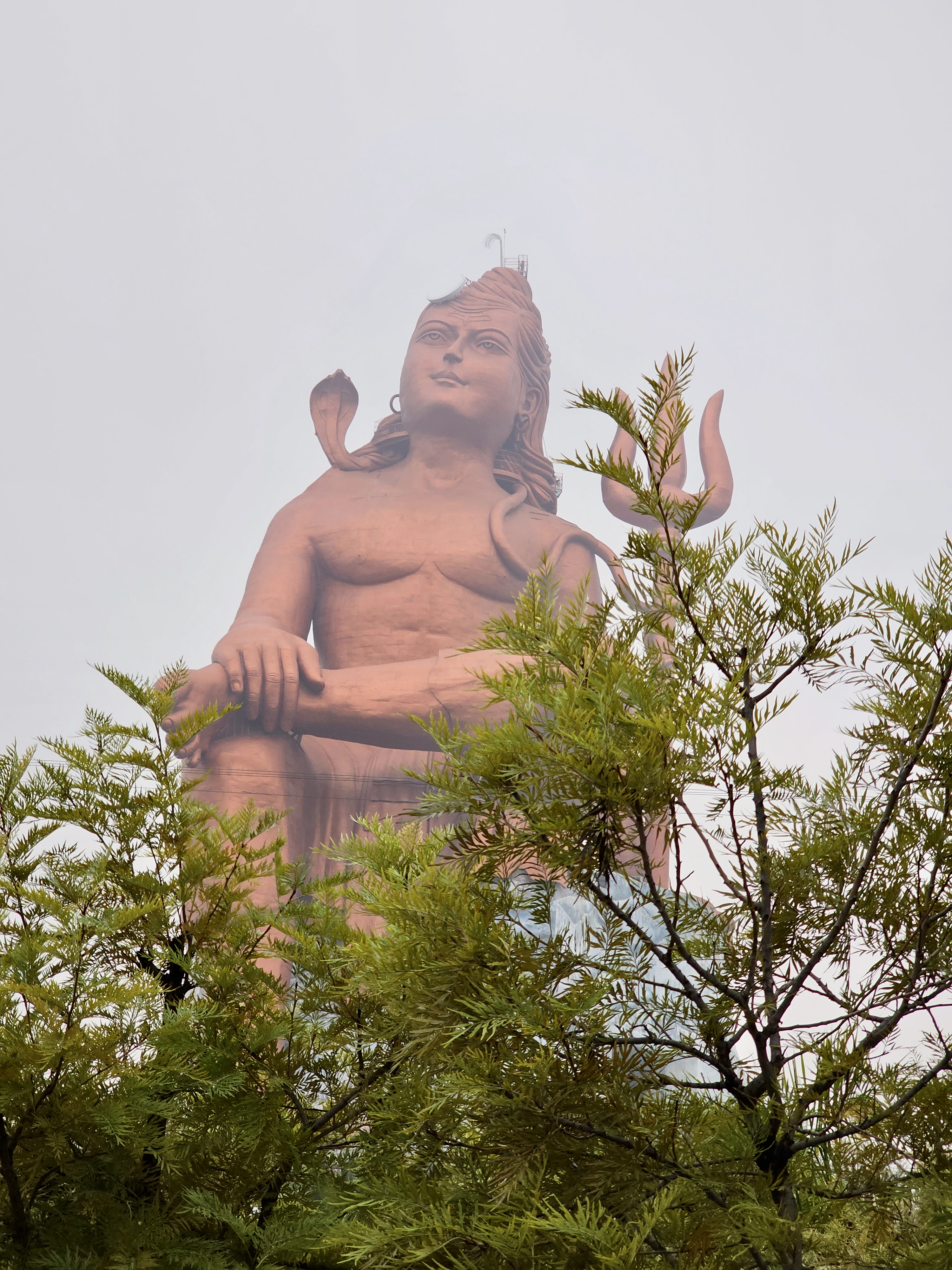 A giant statue of Lord Shiva (Statue of Belief, aka Vishwas Swaroopam) is partially hidden behind green trees and mist, captured near Nathdwara, Rajasthan, on a foggy morning.