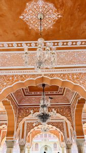 Interior view of Rajasthani palace showing decorative orange walls with white floral patterns, arched doorways, crystal chandeliers, and Mughal-inspired artwork in Jaipur
