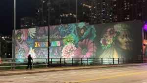 The image is dominated by a building with a painting on the side of flowers and a woman's profile. In the foreground is a street with a woman walking on the sidewalk. It's a night scene. Chicago.