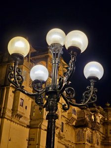 A beautiful, ornate street lamp with five glowing lights stands in the foreground, with the illuminated, historic City Palace of Udaipur visible in the background at night.