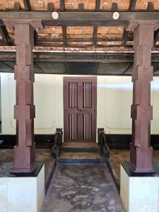 A front view of a heritage entrance inside Hill Palace, with twin wooden pillars and a closed wooden door, set under a traditional tiled roof. Photo taken at Hill Palace, Thrippunithura, Kerala.