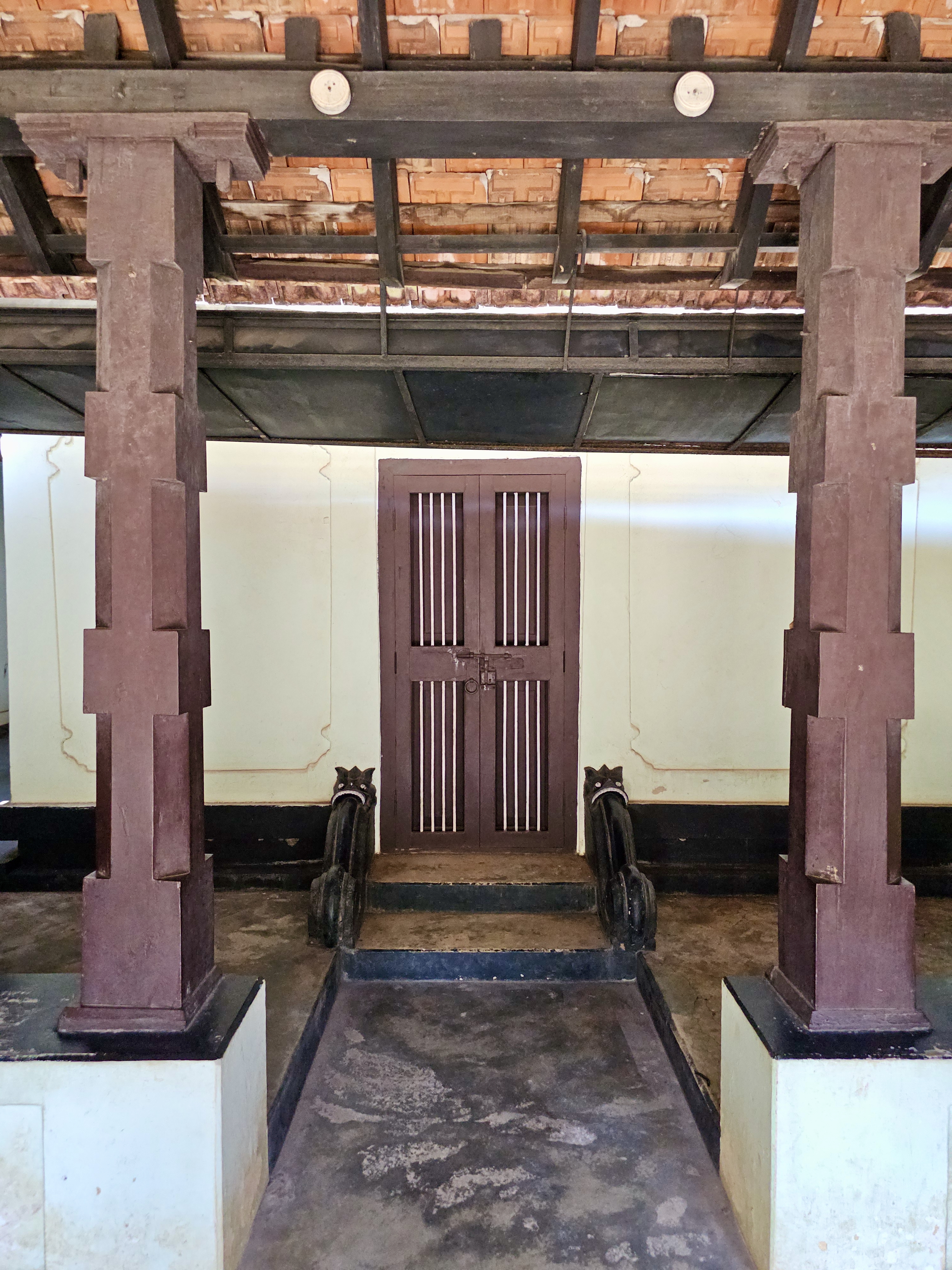 A front view of a heritage entrance inside Hill Palace, with twin wooden pillars and a closed wooden door, set under a traditional tiled roof. Photo taken at Hill Palace, Thrippunithura, Kerala.