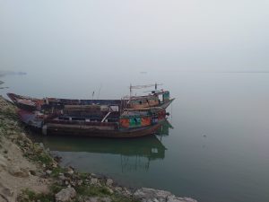 The image features boats on the water, set against a serene backdrop. It captures various types of watercraft, floating peacefully on a lake or river. The scene likely includes elements of nature such as the sky, fog, and surrounding landscapes along the shore. Padma River. Kushtia