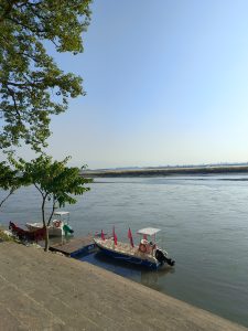 A serene river scene featuring two small boats docked beside a concrete platform. One boat is adorned with red flags, while the other is partially covered with a green tarp.