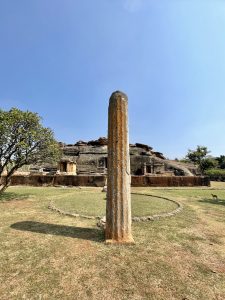 A view of the Ravana Phadi Cave Temple in Aihole, Karnataka, featuring a large stone pillar in the foreground and the rock-cut temple complex behind it.  