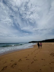 A view of a couple walking nearby beach with sandy ground on the other side, also a view of a cloudy sky is seen.