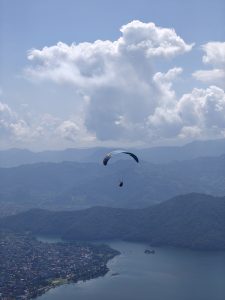 A lone paraglider floats over a town by a shimmering lake, surrounded by green hills under a bright blue sky.