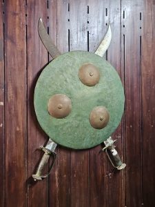 A green iron shield with brass studs and two crossed swords mounted on a wooden wall. This historic weapon display is part of the Hill Palace Museum in Thrippunithura, Kerala.