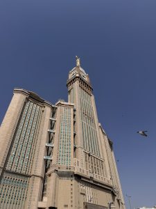 Stunning view of the Abraj Al Bait Clock Tower in Makkah, Saudi Arabia, captured under a clear blue sky with a pigeon flying nearby, showcasing the tower’s golden crescent and detailed Islamic architectural design
