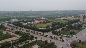 A peaceful landscape at Gulberg Greens, Islamabad — a calm road with just a few cars passing by, surrounded by green farmhouses and open landscape under a soft, cloudy sky.