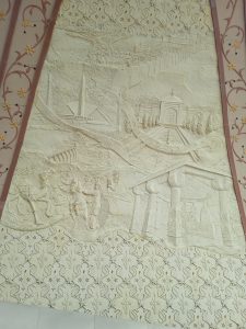 Decorative relief sculpture showing a stylized landscape with hills, a river, towers, and figures on horseback, framed by intricate floral borders