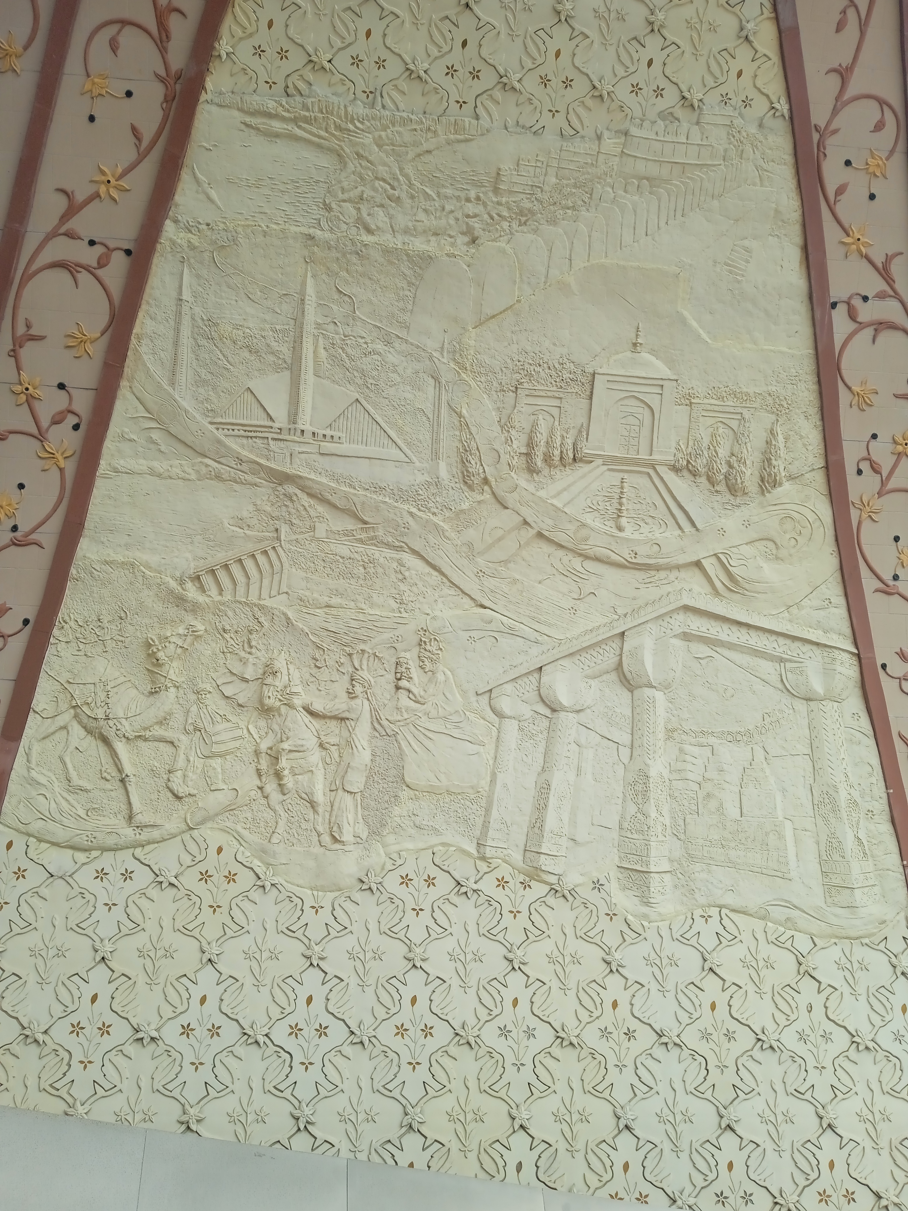 Decorative relief sculpture showing a stylized landscape with hills, a river, towers, and figures on horseback, framed by intricate floral borders