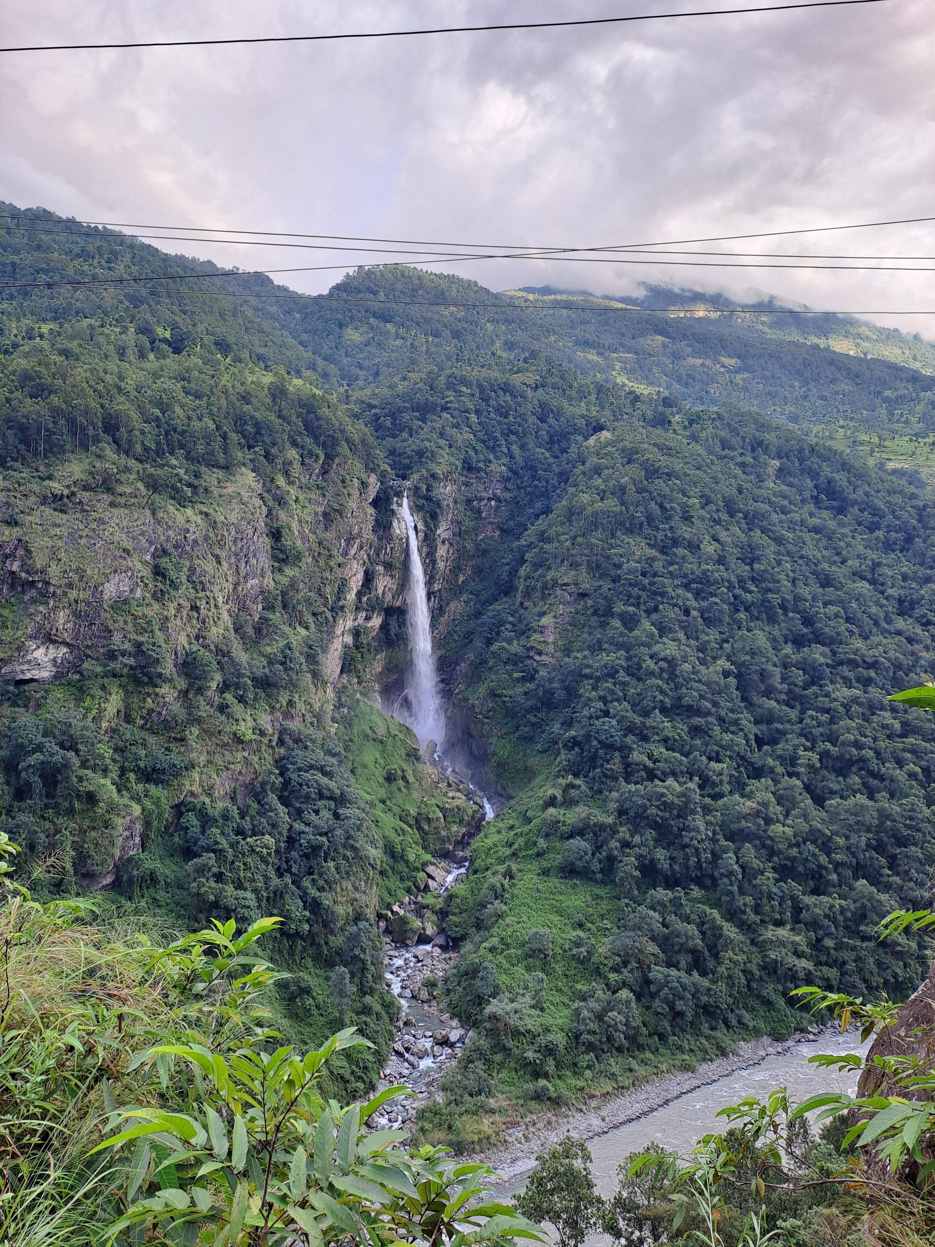 A scenic view of a waterfall cascading down a rocky cliff into a verdant valley, surrounded by lush green hills and dense vegetation. 