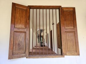 A close-up of a wooden window with vertical iron bars at Hill Palace, Thrippunithura, Kerala. Through the window, a spiral staircase and an interior corridor with arches are visible. 