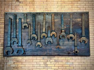 A wall-mounted display of large, antique metal wrenches arranged in two uneven rows on a dark wooden board, set against a tan brick wall. The wrenches vary in size and shape, showing rust and wear, giving the scene an industrial, vintage aesthetic.
