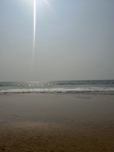 A serene beach scene with gentle waves rolling onto the sandy shore, sunlight reflecting off the water under a hazy sky with a visible sunbeam.