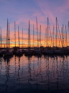 A harbor at sunset. Dark sailboat masts stand tall against a sky of bright orange, pink, and purple. The colorful light is reflected in the water.