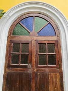 A beautifully crafted old wooden door with small textured glass panels in red, blue, and green colors. The door has a white arched frame and stands out against a bright yellow wall. Captured near Fort Kochi Beach, Kerala. 