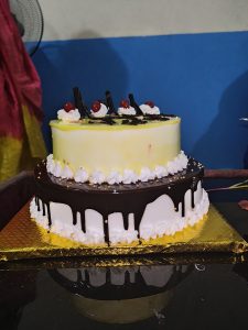 A two-tiered cake featuring a yellow top layer decorated with whipped cream, chocolate shavings, and red cherries. 