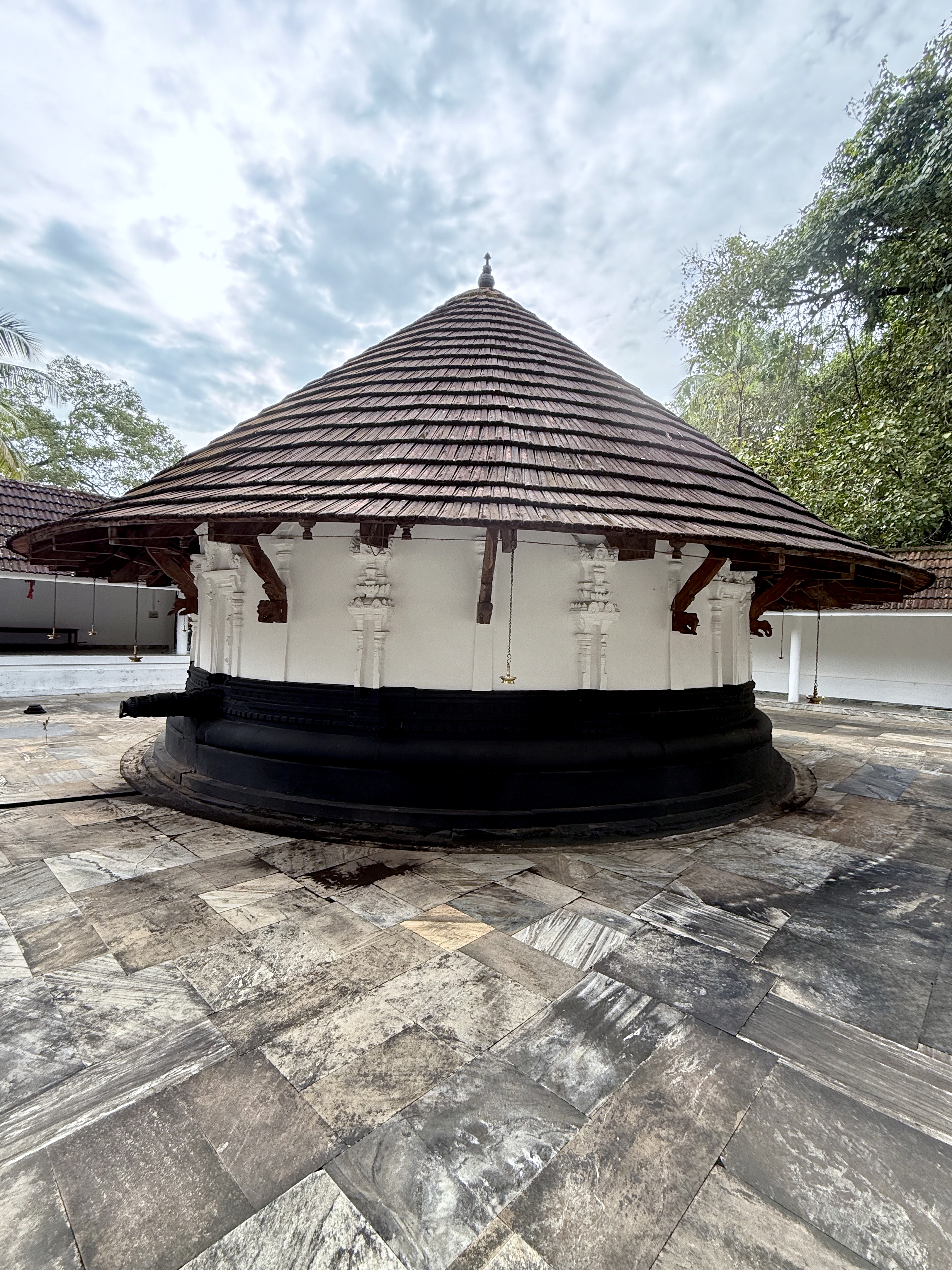 A full view of a round temple sanctum with white walls and a brown wooden-tile roof. The open courtyard is paved with stone slabs, captured from Sree Shiva Vishnu Temple, Perumanna, Kozhikode.