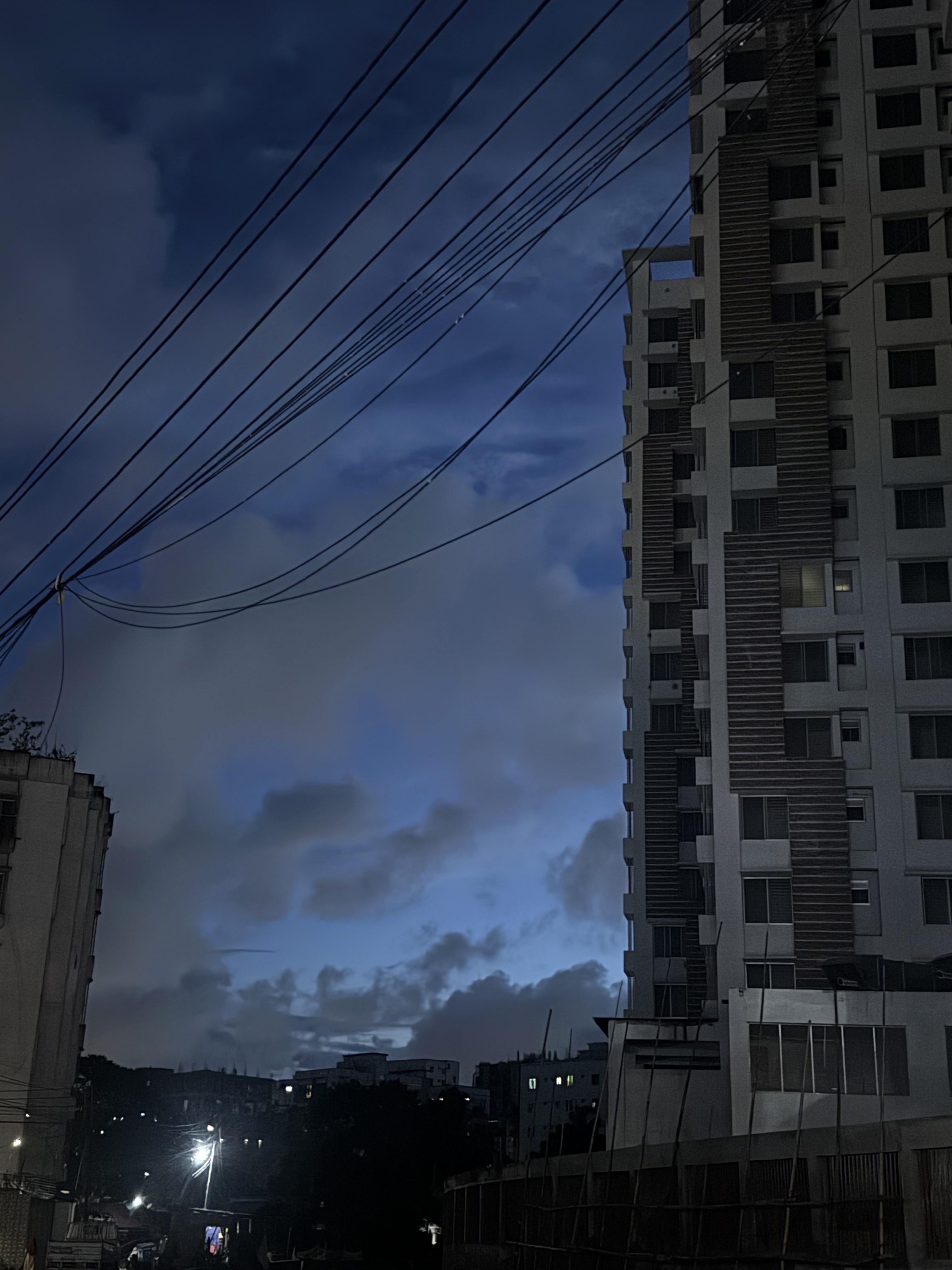 A dusky cityscape with tall apartment buildings and power lines cutting across the frame. The deep blue evening sky with scattered clouds peeks through, while streetlights and windows illuminate the darker streets below.