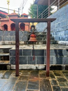 A large temple bell hangs from chains within a dark wooden frame, placed at the bottom of a stone staircase leading up to a red brick structure.