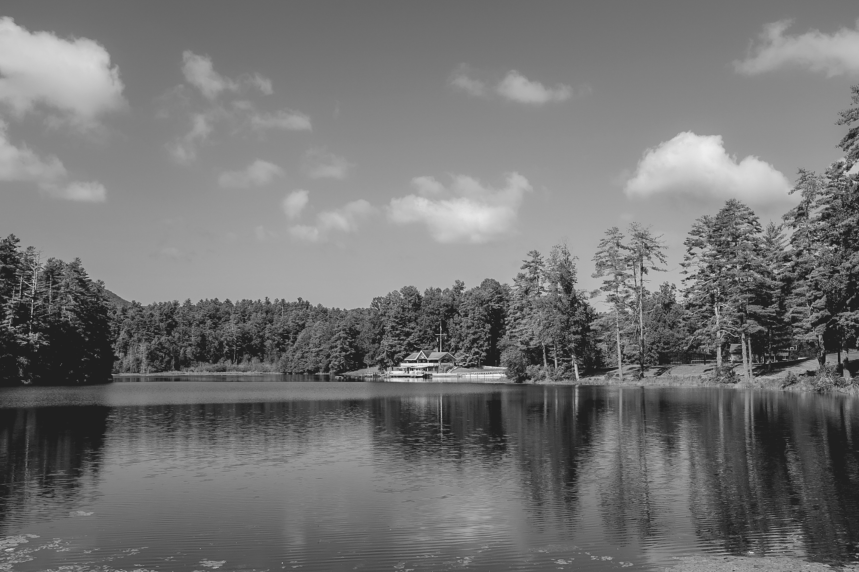 Black and white photo of a lake surrounded by trees with a lodge in the distance and clouds reflecting on the surface.