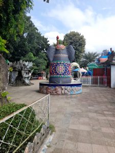 

A colorful, ornate elephant statue with a crown stands in a park with trees, umbrellas, and decorative structures, under a partly cloudy sky.