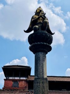 A golden Garuda statue in a prayer pose, sitting atop a stone column with a heritage palace in the background. Photographed at Patan Durbar Square, Lalitpur, Nepal.  