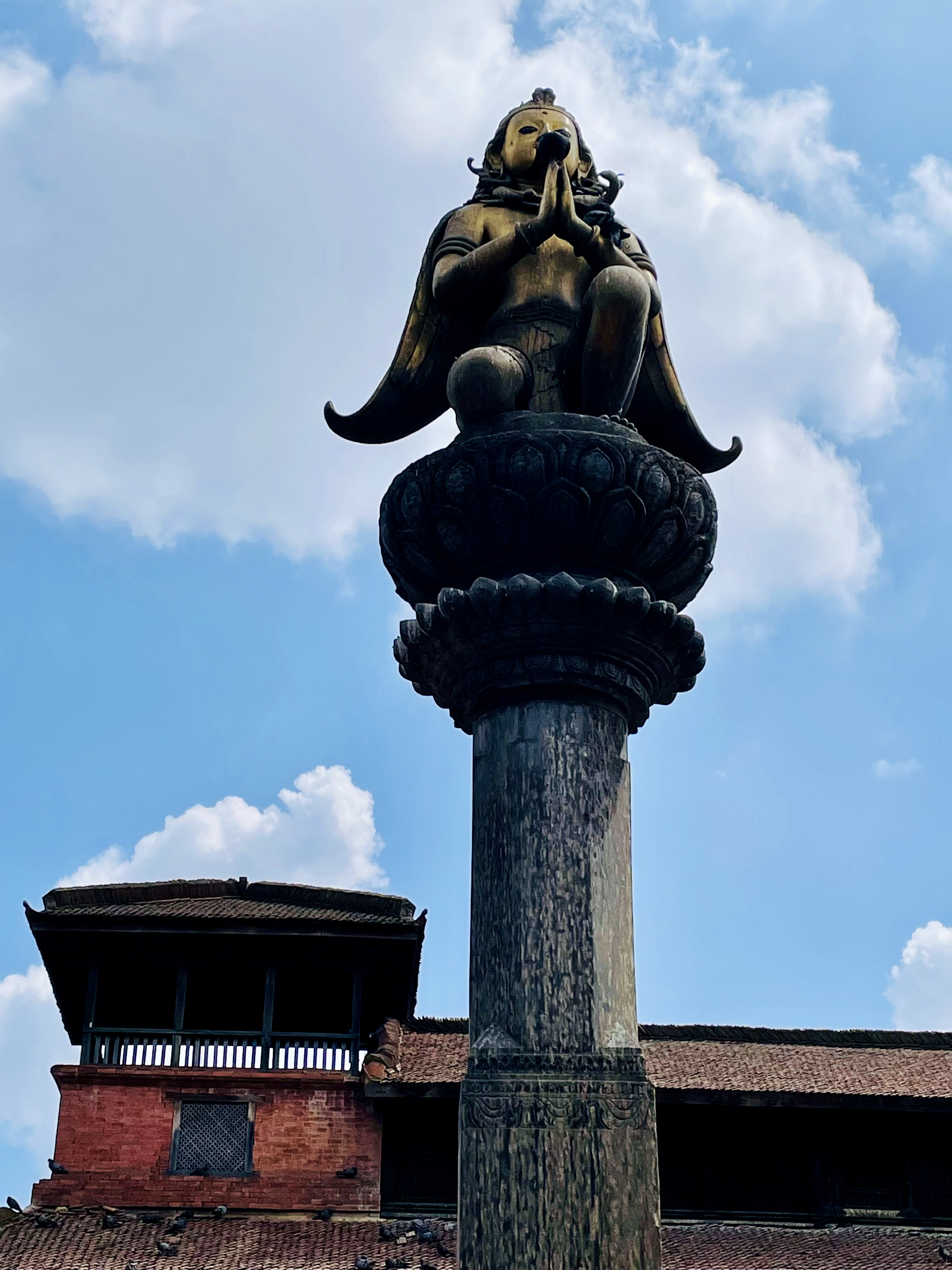 A golden Garuda statue in a prayer pose, sitting atop a stone column with a heritage palace in the background. Photographed at Patan Durbar Square, Lalitpur, Nepal.
