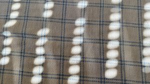 A textured surface featuring a soft fabric in light beige with a plaid pattern. The fabric is illuminated by sunlight, casting a series of circular light and shadow patterns across the material, creating a play of light and texture.