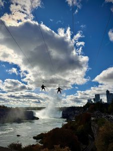 Two people ziplining high above Niagara Falls on parallel cables, silhouetted against a bright blue sky with dramatic clouds. Below them, mist rises from the powerful waterfall, while the city skyline and observation areas line the riverbank surrounded by autumn-colored trees.
